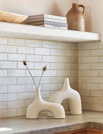 #color::white #size::small #size::large | The Leonor sculptural arched matte white ceramic Vase in its small and large size sitting atop a granite countertop in a kitchen with a subway tile backsplash, a stack of books, and a wooden centerpiece bowl