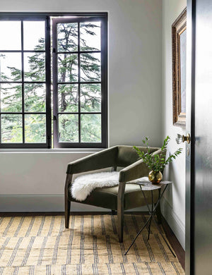 The Lyssa olive velvet accent chair sits in the corner of a room with a striped rug and a white sheepskin blanket thrown atop it.