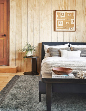 The Illy metal geometric side table sits in a bedroom with a gray linen bed, a gray plush rug, and a light wood paneled wall