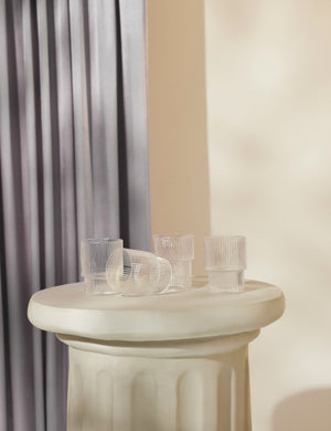 The Art deco inspired rian ripple set of four clear glassware sit on a cream pedestal with a gray curtain draped in the background