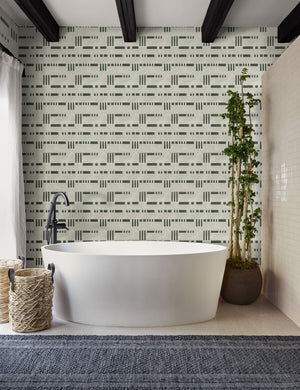 The Artemis green and white dash-and-bar motif patterned wallpaper is in a bathroom with a white oval bathtub, a slate gray runner rug, and a natural tiled wall