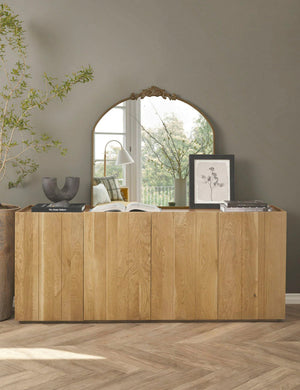 The Tulca arched gold mirror with flat bottom edge and traditional scroll detailing sits atop a wooden side table with a sculptural vase and floral wall art.
