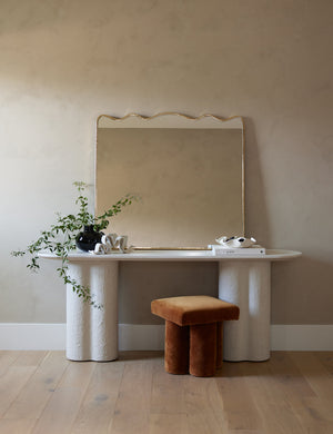  The rook mantel mirror sits atop a white sideboard with pedestal legs next to a cognac velvet ottoman