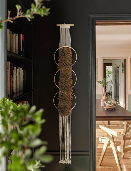 #showall::39842068660323 | The Studio Nom Lwazi light brown woven Wall Hanging by nom hangs in a black painted room next to a bookshelf and a dining room in the background