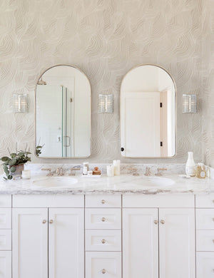 Alina neutral Wallpaper with smooth ripple pattern is in a bathroom with white countertops and cabinetry and two arched mirrors