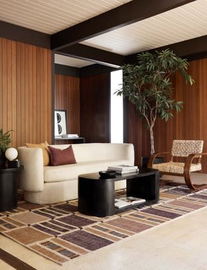 The Luna black wood oval coffee table sits in a retro living room atop a burgundy and orange patterned rug surrounded by a cream boucle rounded couch and a woven accent chair with wooden arms and legs.