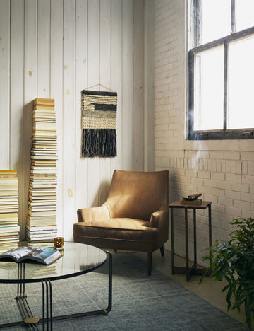 | The Ilona Leather Accent Chair sits in the corner of a living room next to a round coffee table and two high stacks of books