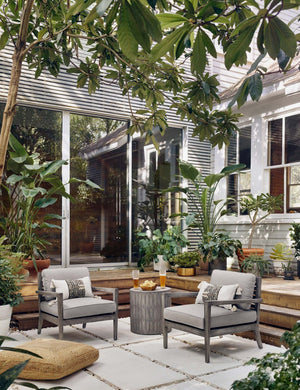 The Glyn concrete indoor and outdoor side table with carved finish sits in an outdoor space between two gray wooden framed accent chairs atop a tiled concrete floor