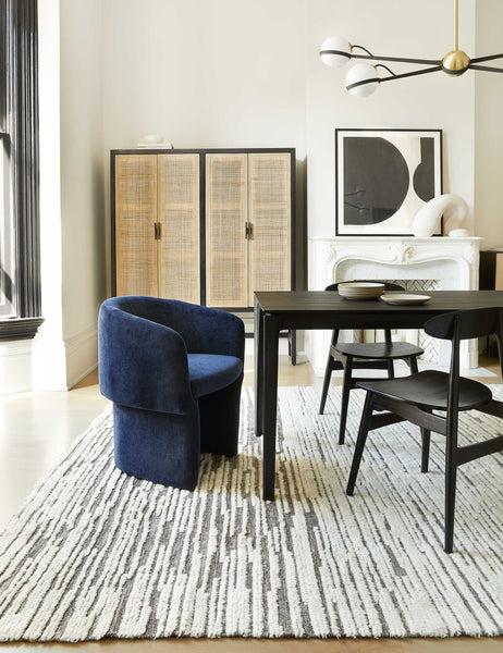 | The Adeline black and brass retro chandelier sits in a dining room with a black wooden dining table, black wooden dining chairs, and a navy velvet accent chair