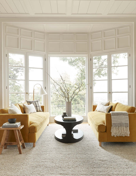 #showall::39490333343843 | The Elulia woven two-tone taupe throw with fringed ends lays atop a yellow velvet sofa in a living room with an oval coffee table, white accented walls, and floor to ceiling french doors and windows