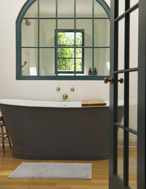 The Cloud loom sustainable light gray bath mat by coyuchi lays on a hardwood floor in a bathroom with a black bathtub and an arched mirror