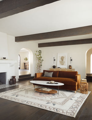 The Zehra hand-knotted ivory, gray and purple medallion wool-blend area rug lays in front of a rust colored sofa and under an oval coffee table with a white top and wooden shelf.