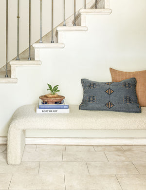 The Indigo woven cotton boho and folk-inspired lumbar pillow with a wool front sits on a white boucle bench with a stack of books and an orange throw pillow