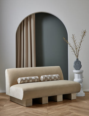  The Lena armless brie beige velvet sofa sits in a studio with two checkerboard bolster pillows atop it