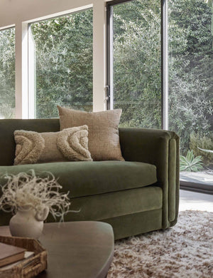 The arches ivory lumbar pillow sits atop a green velvet sofa in a room with floor to ceiling windows