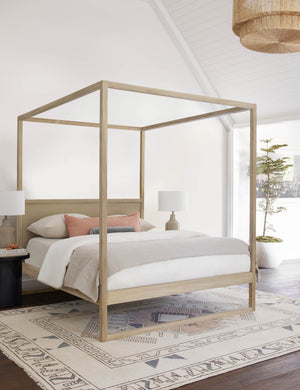 The Kiery light wood canopy bed sits in a bright bedroom with a sloped ceiling, neutral colored bedding, and a multicolored patterned rug.