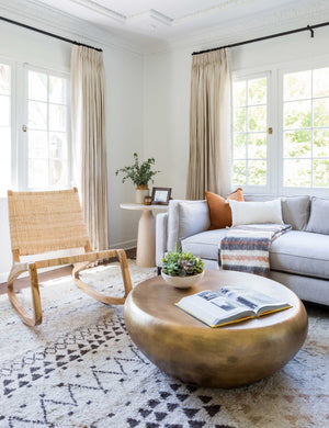 The Arteriors bates round metallic coffee table sits in a living room with a woven accent chair, a gray linen sofa, and a geometric patterned rug.