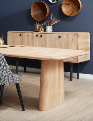 The Mela oak wood dining table sits in a dining room with a white and black patterned chair and an oak wood side table with four cabinets below wooden wall hangings.
