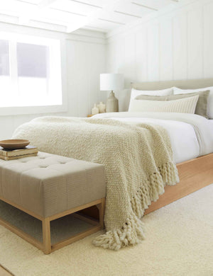 The Olema ivory handwoven throw with fringed ends lays in a bedroom with a cream plush rug, a wooden-base bed, and a beige cushioned bench
